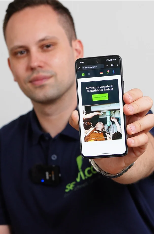 Provider in a navy servi.co polo holding a phone showing the servi.co mobile home with a prominent search-services button in German.
