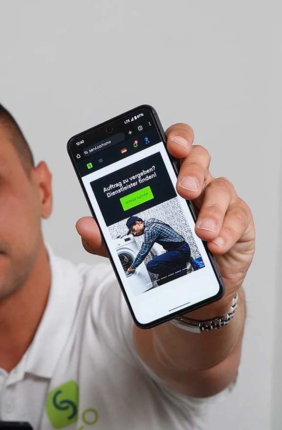 Service provider in a white polo holding a phone showing the servi.co home hero and search service CTA in German.